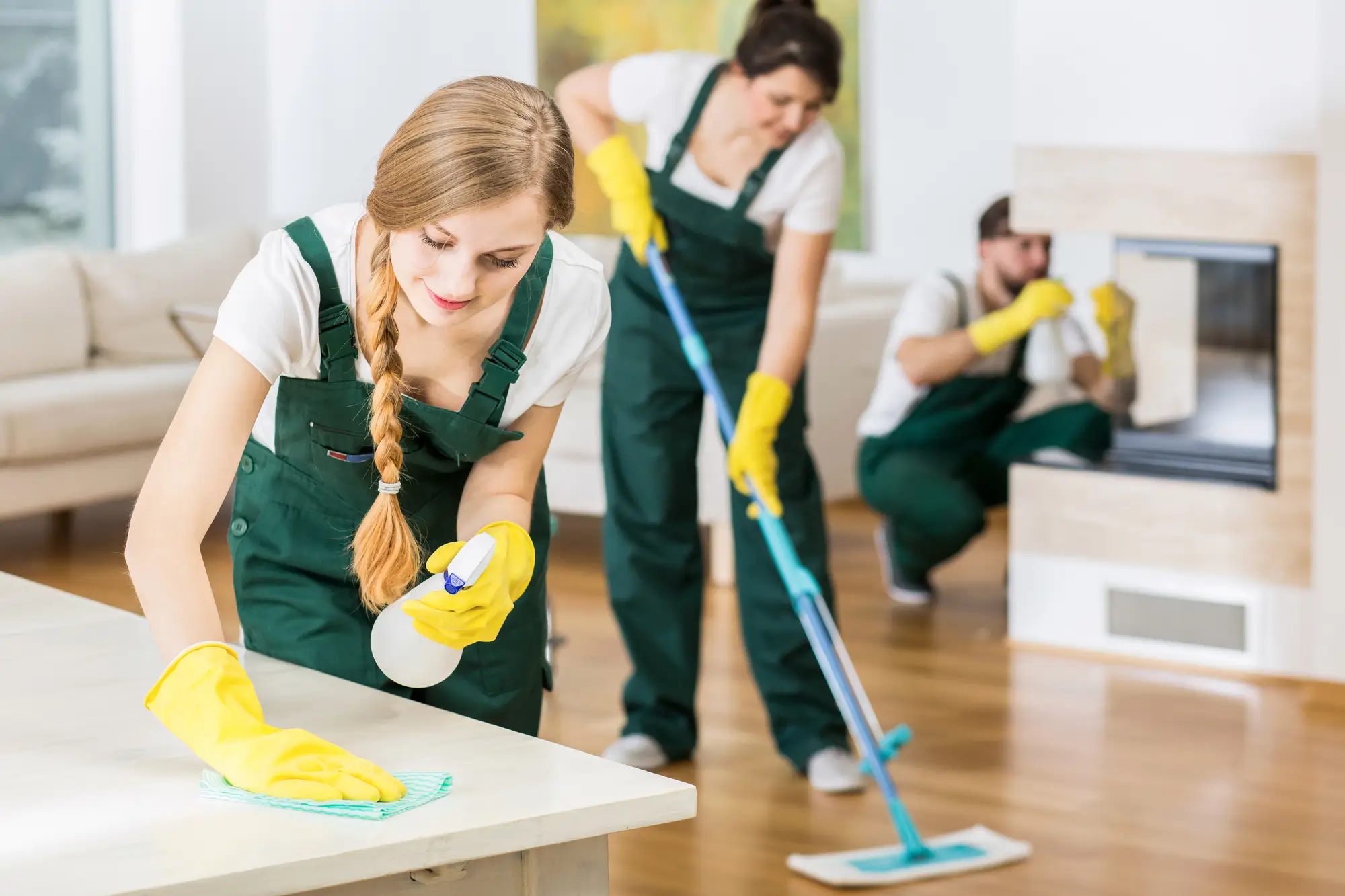 Professional cleaners in uniform disinfecting table, mopping floor, and cleaning fireplace